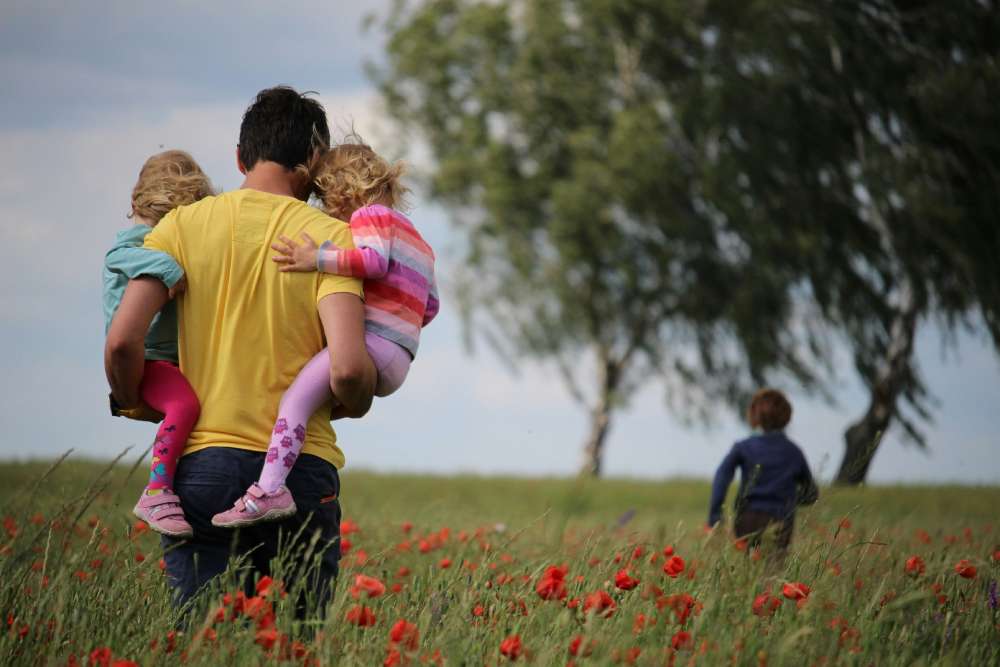 A father carries his children while walking through a poppy field on a sunny day