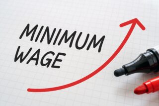 A white boxed background with minimum wage written on it with black ink and a red upward arrow