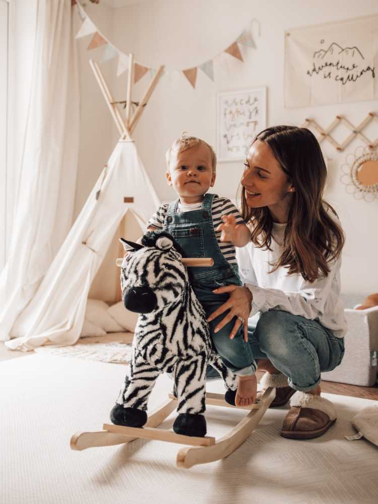 A cheerful mother watches over her toddler as he sits on a rocking horse